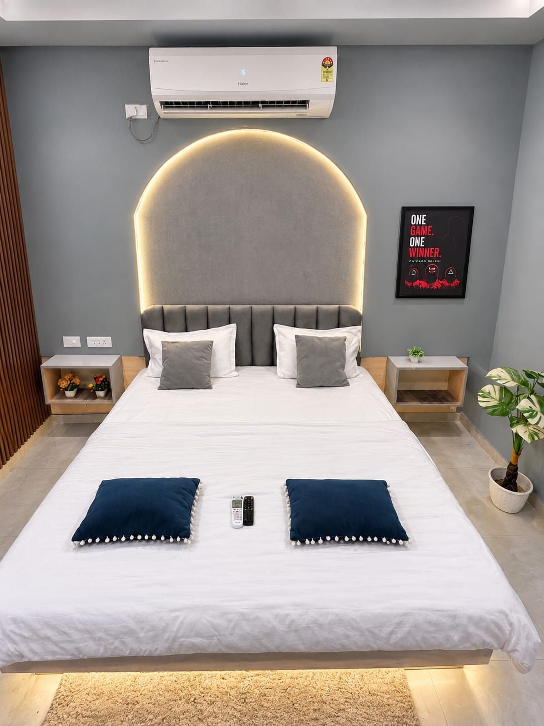 Quarto moderno com uma cama grande, mesas de cabeceira, ar condicionado fixado na parede, um pôster emoldurado e uma planta em vaso.