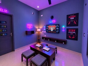A room with a neon sign reading 'EL 01', a television displaying 'TEKKEN 3', and a wall with 'GAME ON' and 'STAY IN THE GAME' posters. - The Arcade Stay (Greater Noida)