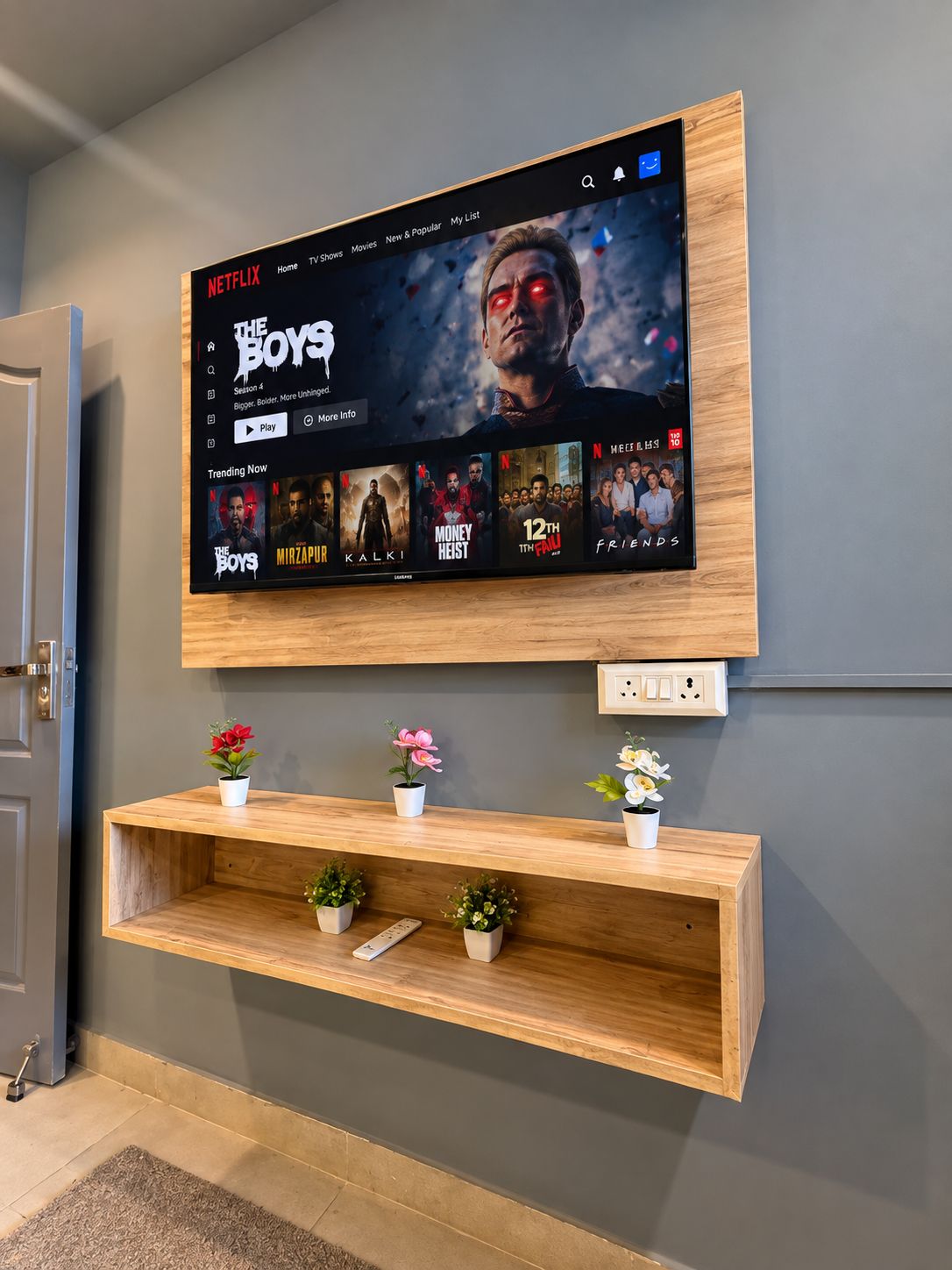 A flat-screen TV mounted on a wall displaying the Netflix interface with various shows. Below the TV, there is a wooden shelf with potted plants.