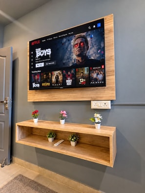 A flat-screen TV mounted on a wall displaying the Netflix interface with various shows. Below the TV, there is a wooden shelf with potted plants. - The Arcade Stay (Greater Noida)