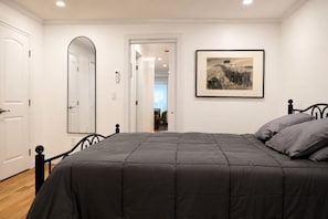 Ein Schlafzimmer mit einem Bett, einer Tür, einem Spiegel, einem gerahmten Bild an der Wand und einem Flur, der zu einem anderen Raum führt.