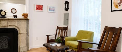 A living room with a fireplace, two wooden chairs, a small table with a plant, and artwork on the walls.