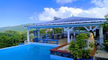 A pool area with a covered seating area, a view of the surrounding landscape, and a clear sky.