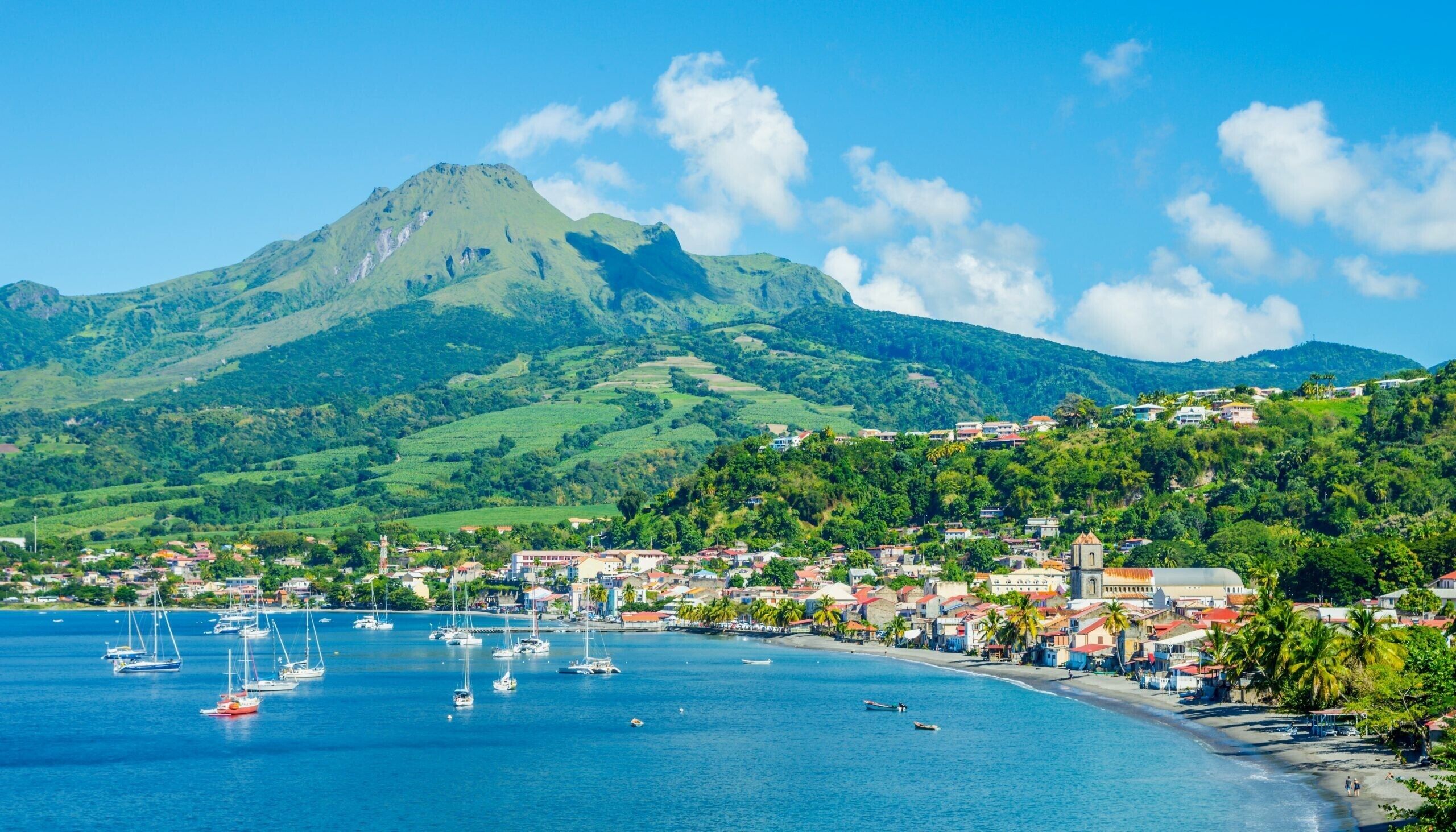 Une ville côtière, avec une montagne en arrière-plan, des bateaux sur l’eau et des bâtiments le long de la côte.