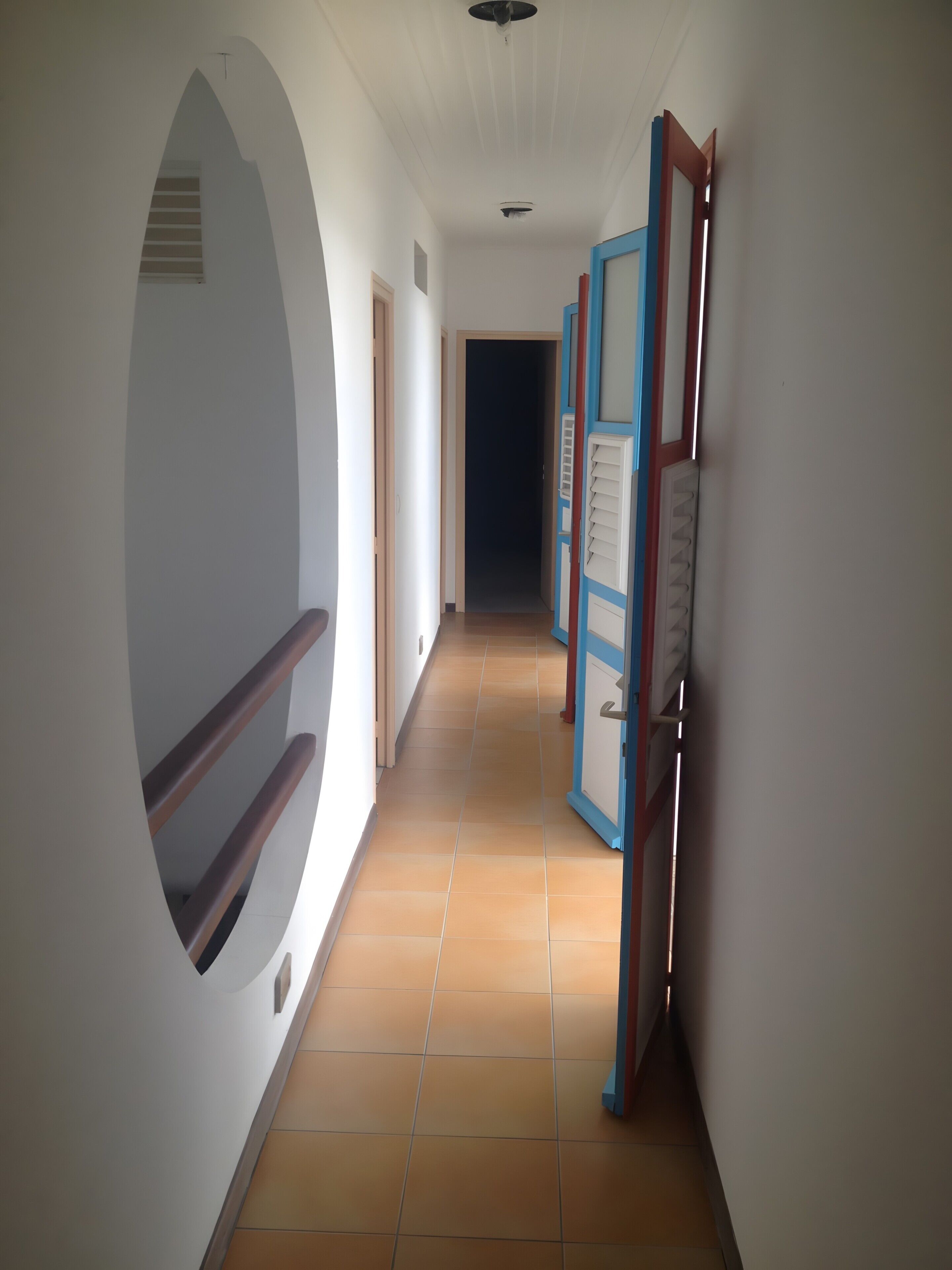 Un couloir avec une fenêtre circulaire, une porte dotée d’un cadre bleu et un sol carrelé.