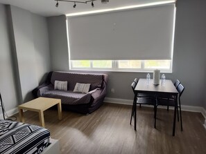 Um quarto com sofá cinza, mesa de centro de madeira, mesa de jantar com cadeiras e uma janela com persianas.