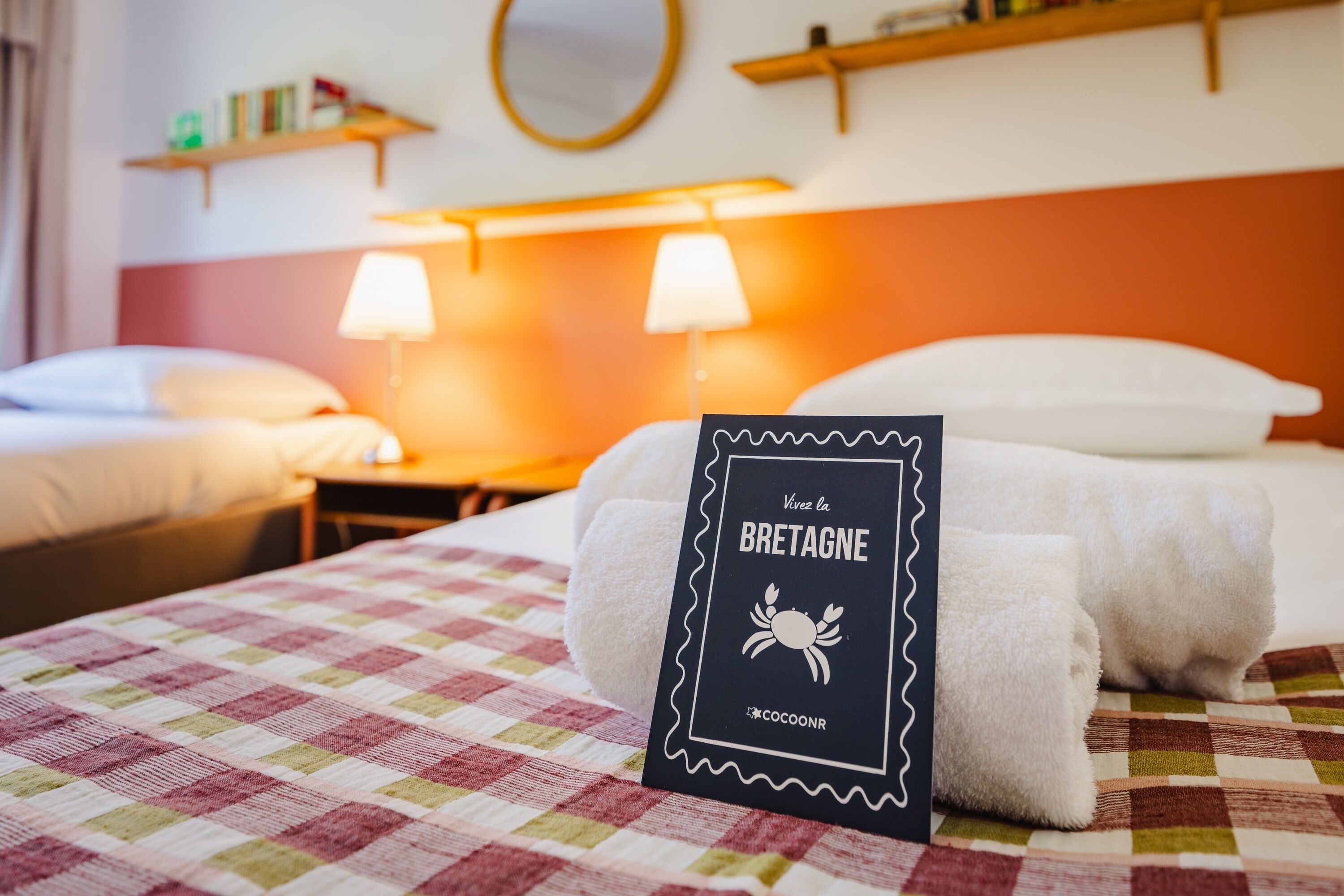 Een hotelkamer met een geruit sprei, een bordje met de tekst 'Vivez la BRETAGNE' op het bed, en een nachtkastje met een lamp.