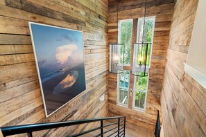 A wooden interior with a large framed picture of a beach scene, a staircase with a metal railing, and a window with a view of greenery. - Luxury on Eastern Lake | Lakefront | Private Pool | 6 Beach Bikes (Santa Rosa Beach)