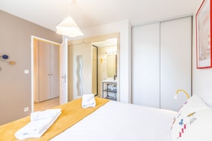 A hotel room with a bed, a wooden headboard, a bedside table with towels, a wardrobe, a bathroom with a mirror, and a framed picture on the wall. - Capsea - Vue sur le port (Capbreton)
