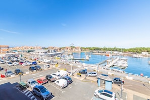A marina with boats docked, a parking lot with cars, and buildings in the background. - Capsea - Vue sur le port (Capbreton)