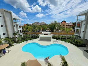 A swimming pool with a curved edge, surrounded by a white building with balconies, palm trees, and a clear blue sky. - Laguna Beach 306 Stylish Apartment- Just 3 Minutes Walking to Playa Dominicus (Dominicus)