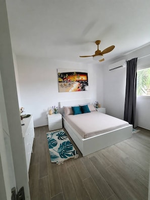 A bedroom with a bed, nightstands, a ceiling fan, a painting, and a window with curtains. - Laguna Beach 306 Stylish Apartment- Just 3 Minutes Walking to Playa Dominicus (Dominicus)
