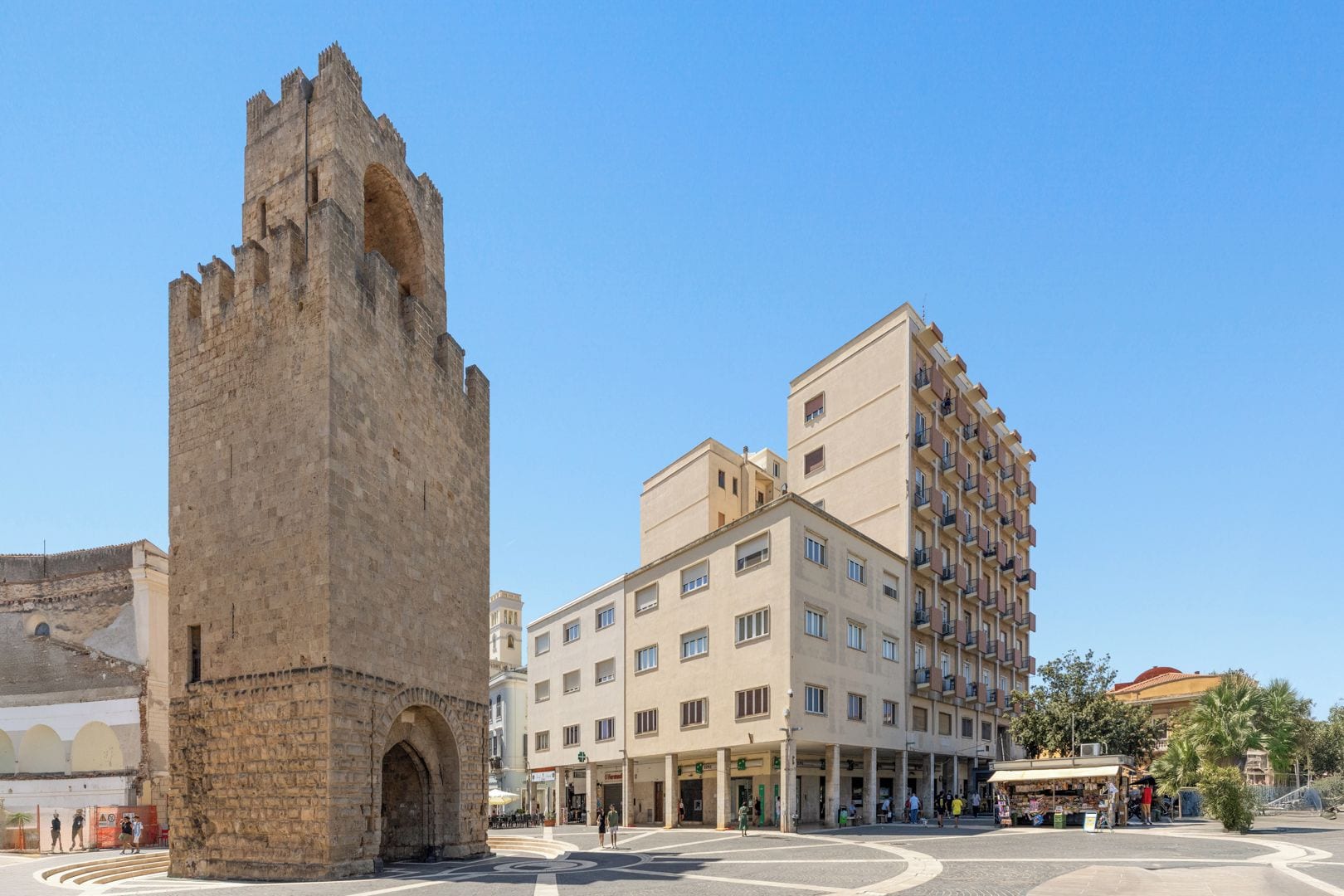 Una torre de piedra histórica y un moderno edificio de varios pisos con balcones, ubicados en una plaza pública.