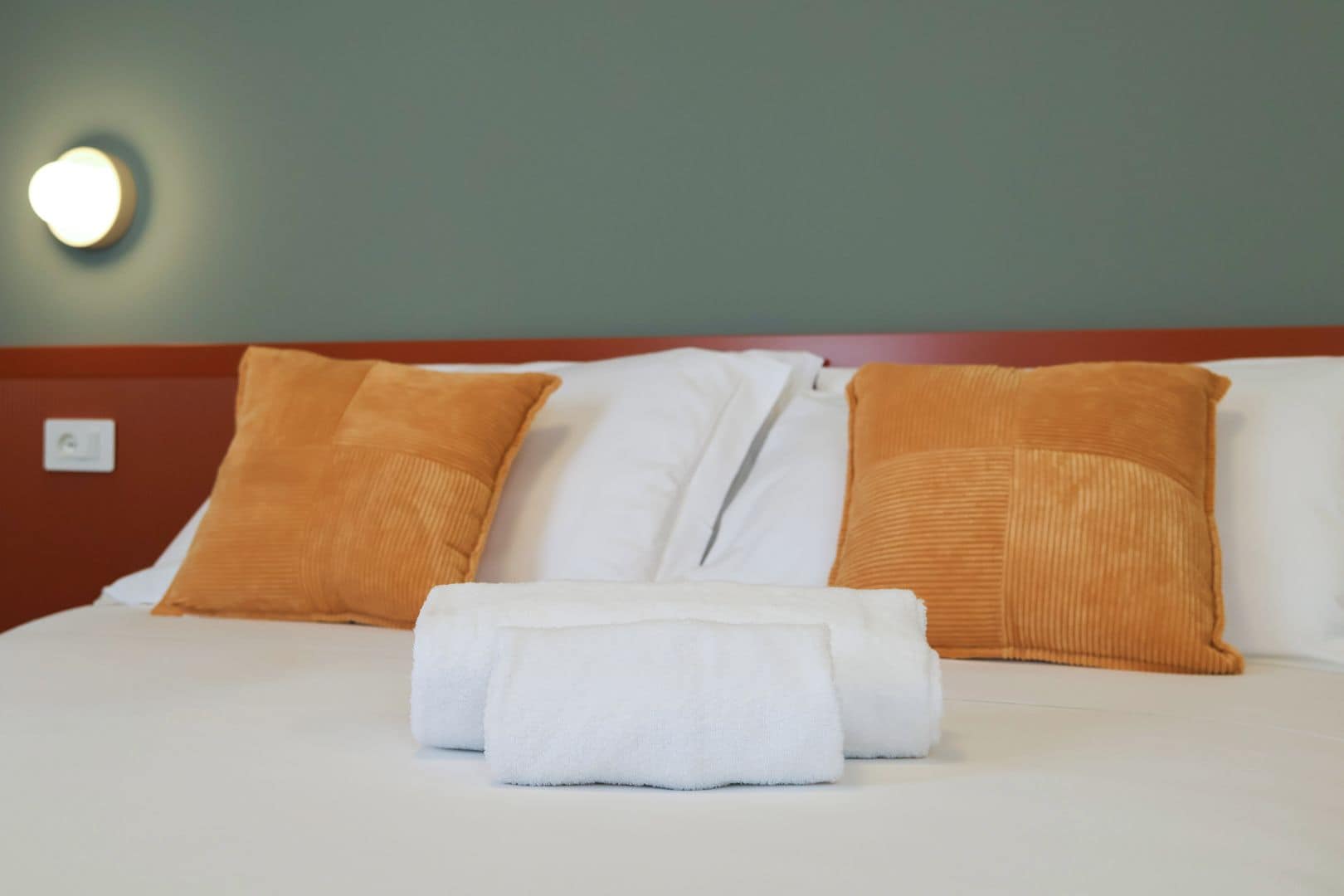 Cama bem arrumada com lençóis brancos e duas almofadas laranjas, uma toalha branca e luminária fixada na parede.
