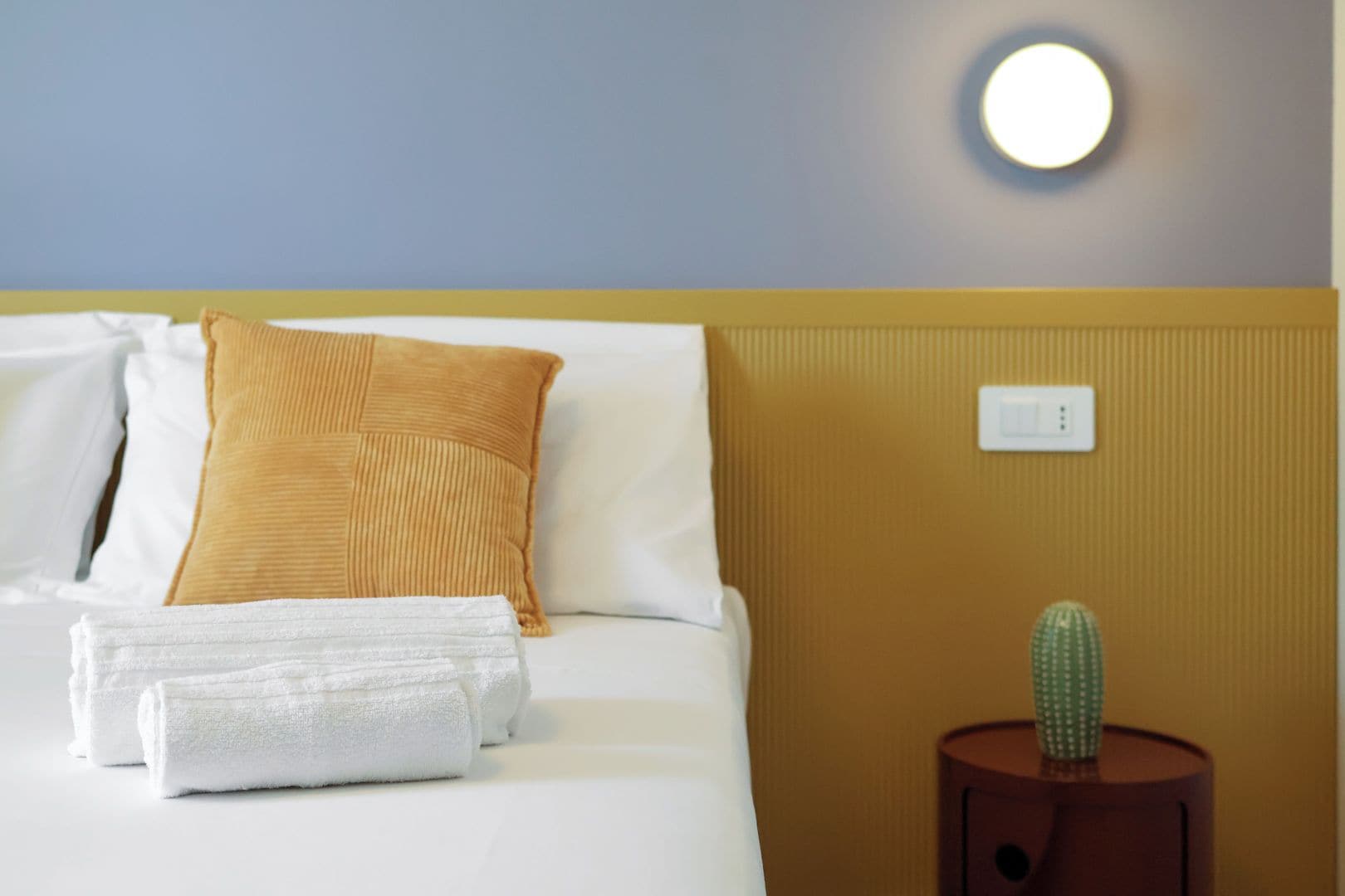Una camera d'albergo con un letto, un cuscino, un asciugamano bianco, un comodino con un cactus e una lampada fissata al muro.
