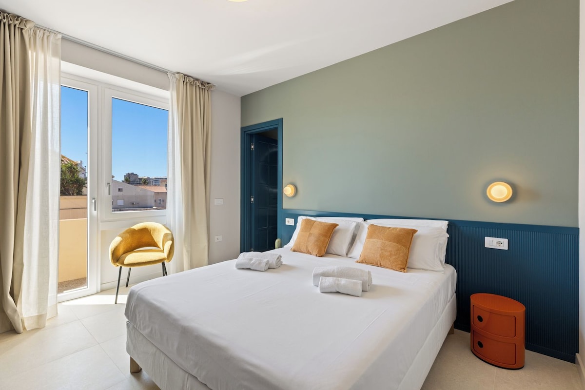 A hotel room with a bed, a chair, a nightstand, and a view through a window.