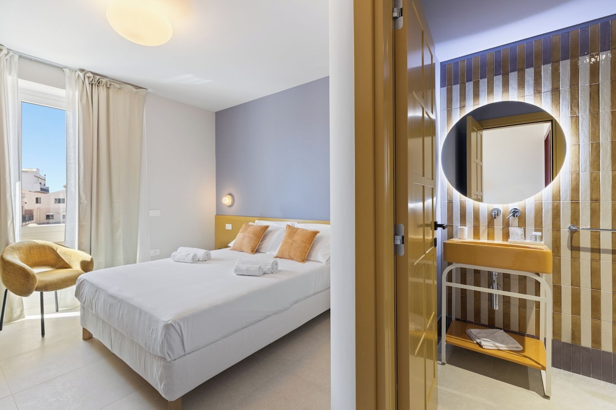A hotel room with a bed, a chair, a bathroom with a mirror, and a view of the outside.
