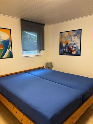 Une chambre avec un lit simple, une fenêtre avec un store et deux tableaux accrochés aux murs.