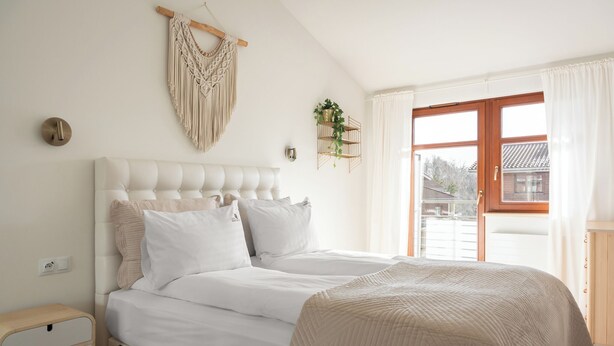 Quarto com cabeceira estofada, duas camas com roupa de cama branca, mesa de cabeceira, peça de macramê na parede e janela com cortinas.
