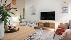 Uma sala de estar moderna com uma mesa de jantar de madeira, cadeiras brancas, um sofá, uma TV e obras de arte nas paredes.