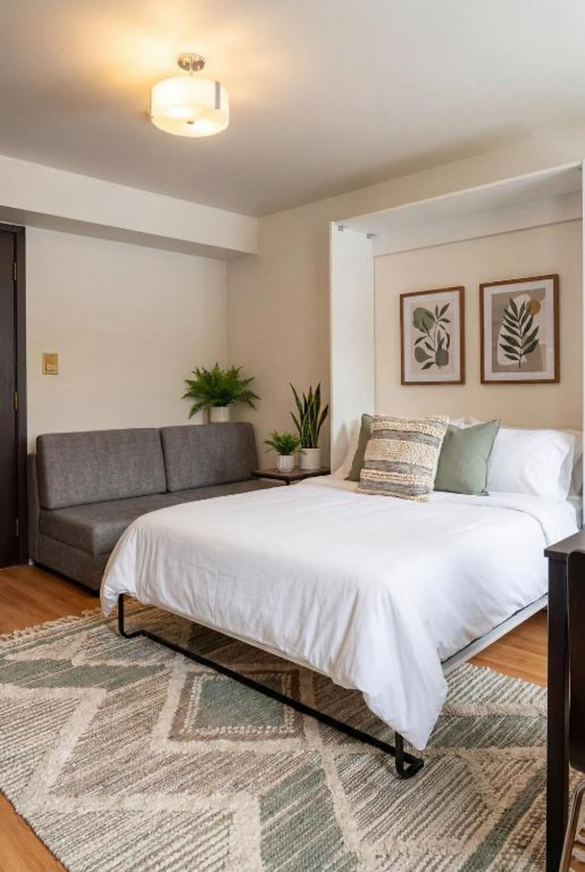 A bedroom with a bed, a grey sofa, a small potted plant, and two framed leaf artworks on the wall.