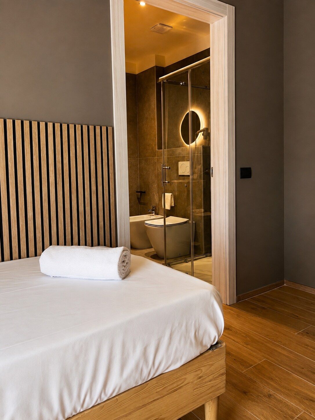 A modern hotel room with a wooden headboard, a neatly made bed, and a glass-enclosed shower area.