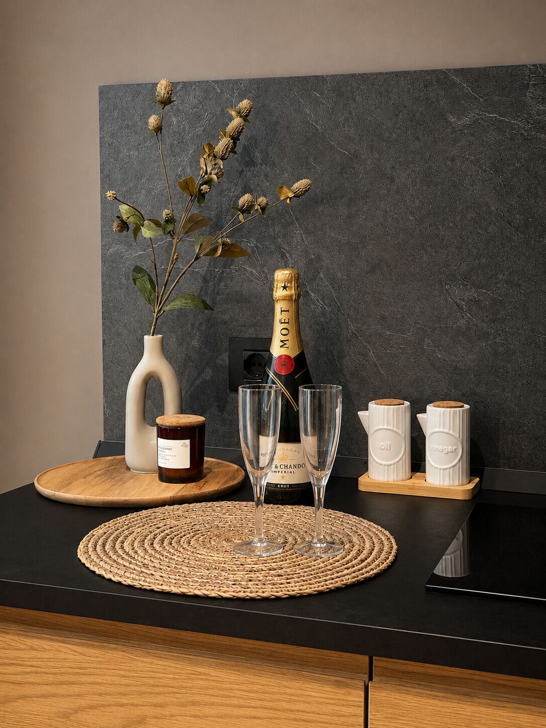 Une table avec une bouteille de champagne, deux verres, un vase contenant des fleurs séchées et deux récipients étiquetés « huile » et « vinaigre ».