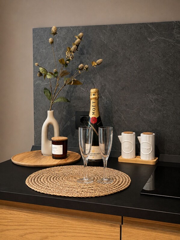 A table with a bottle of champagne, two glasses, a vase with dried flowers, and two containers labeled 'oil' and 'vinegar'.