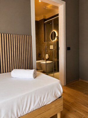 A hotel room with a bed, a wooden headboard, a glass-enclosed shower, and a visible bathroom with a toilet and sink.