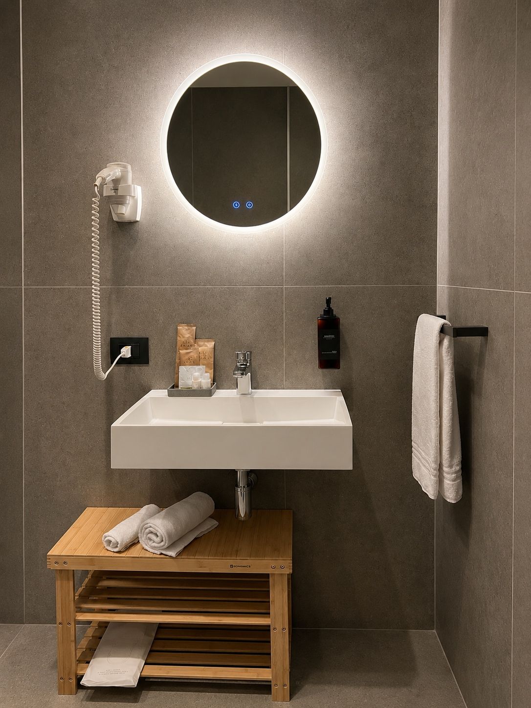 Un bagno moderno con un lavandino bianco, uno specchio rotondo con illuminazione integrata, un porta asciugamani e uno sgabello in legno.