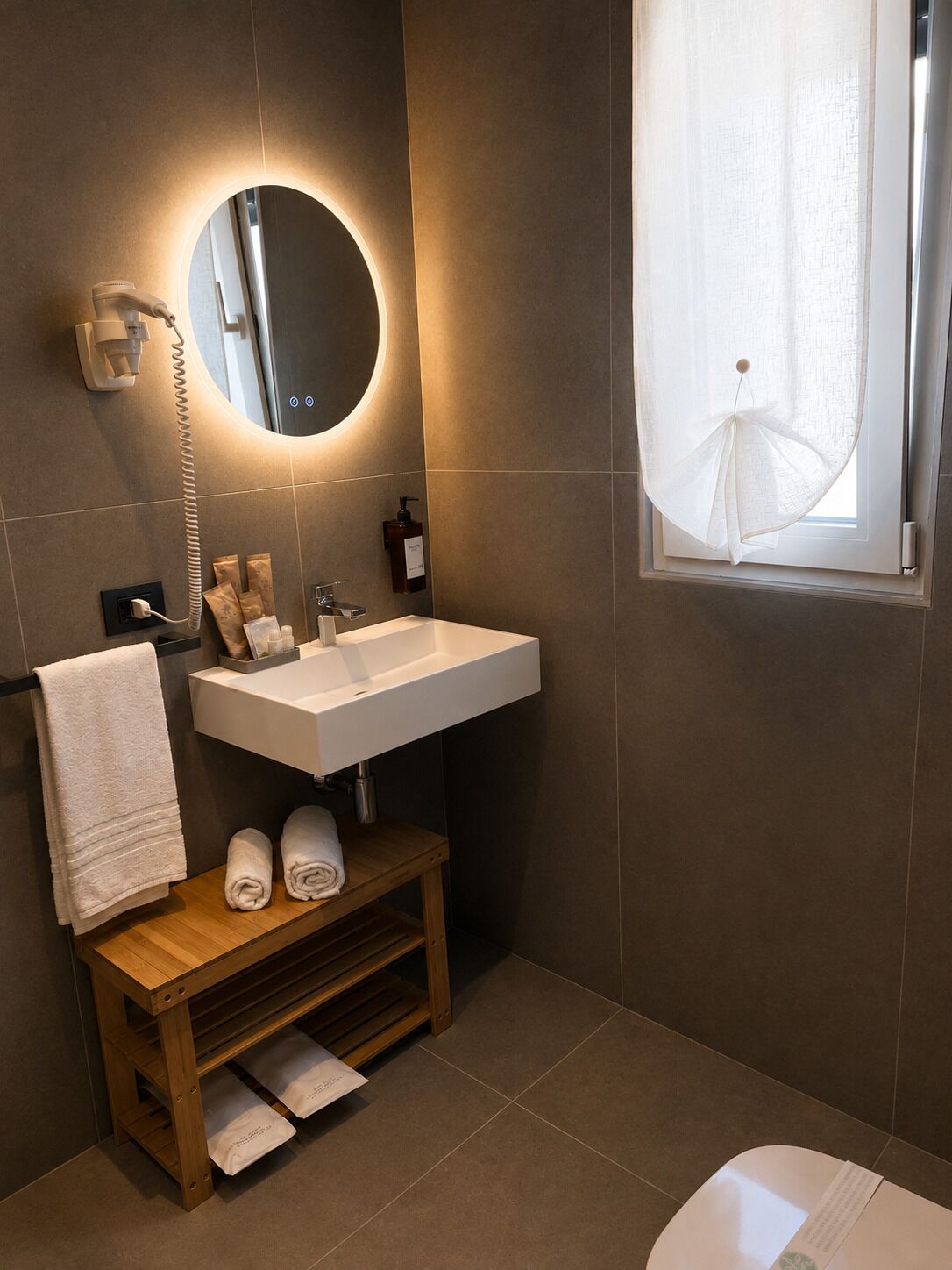 Un baño moderno con un lavamanos blanco, un espejo redondo, un perchero para toallas y una ventana con cortina translúcida.