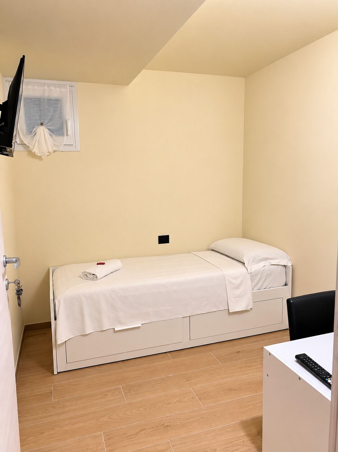 Une petite chambre avec un lit simple, un plancher en bois, un lit blanc, un petit bureau avec une télécommande et une fenêtre avec un rideau.
