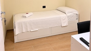 Une petite chambre avec un lit simple, un plancher en bois, un lit blanc, un petit bureau avec une télécommande et une fenêtre avec un rideau.