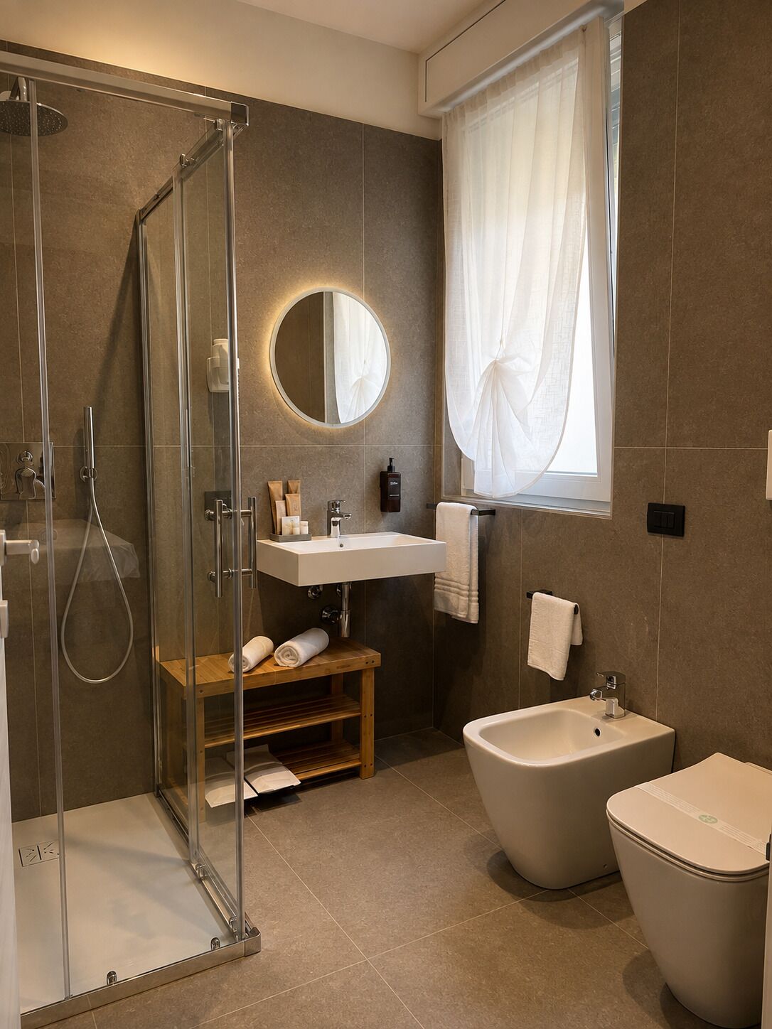 Un baño moderno con ducha envuelta en vidrio, un espejo redondo, un lavamanos, un lavavolos y un inodoro.