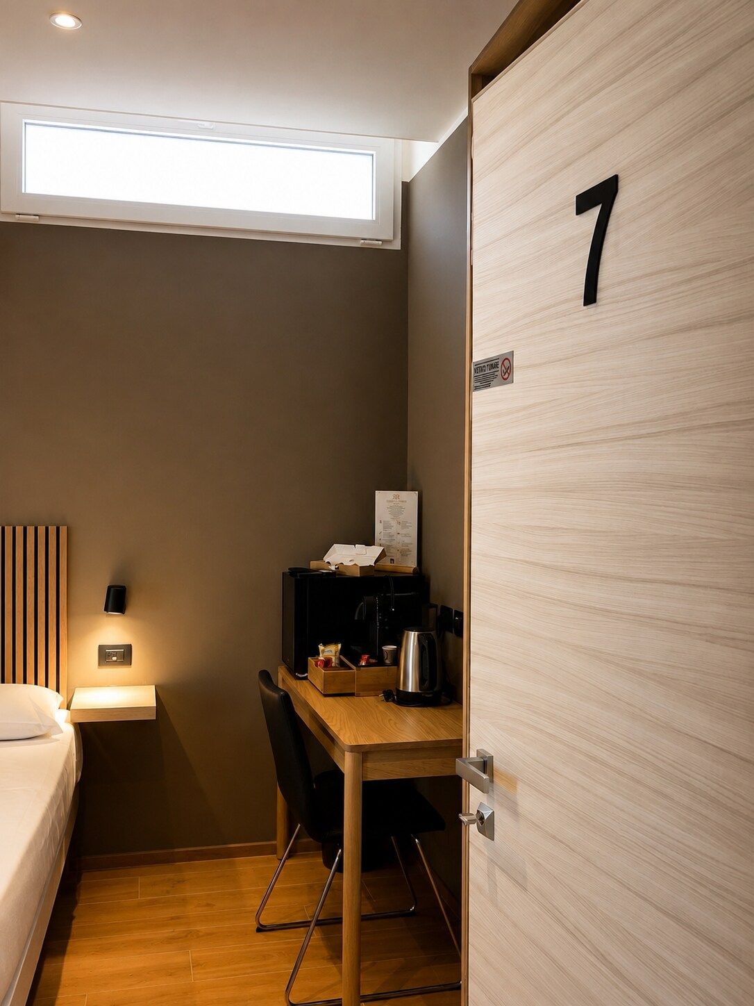 Una habitación de hotel compacta con una cama, un escritorio con una silla, una cafetera y una mesita pequeña. Se ve una puerta con el número 7.