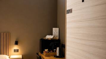Una habitación de hotel compacta con una cama, un escritorio con una silla, una cafetera y una mesita pequeña. Se ve una puerta con el número 7.