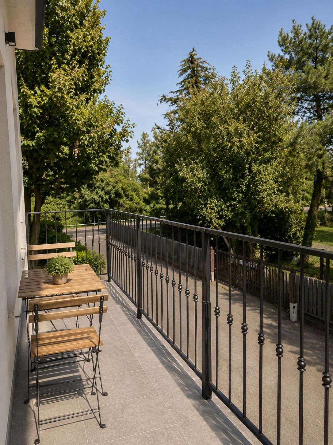 Un balcon avec une table et des chaises en bois, un pot de fleurs et une rambarde en métal donnant sur une rue bordée d’arbres.