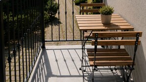 Een balkon met een houten tafel en bankjes, een bloempot en uitzicht op een straat met bomen en gebouwen.