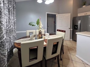 A dining area with a round table, chairs, a vase with flowers, and a kitchen with a refrigerator in the background. - Amos Dr (Greensboro)