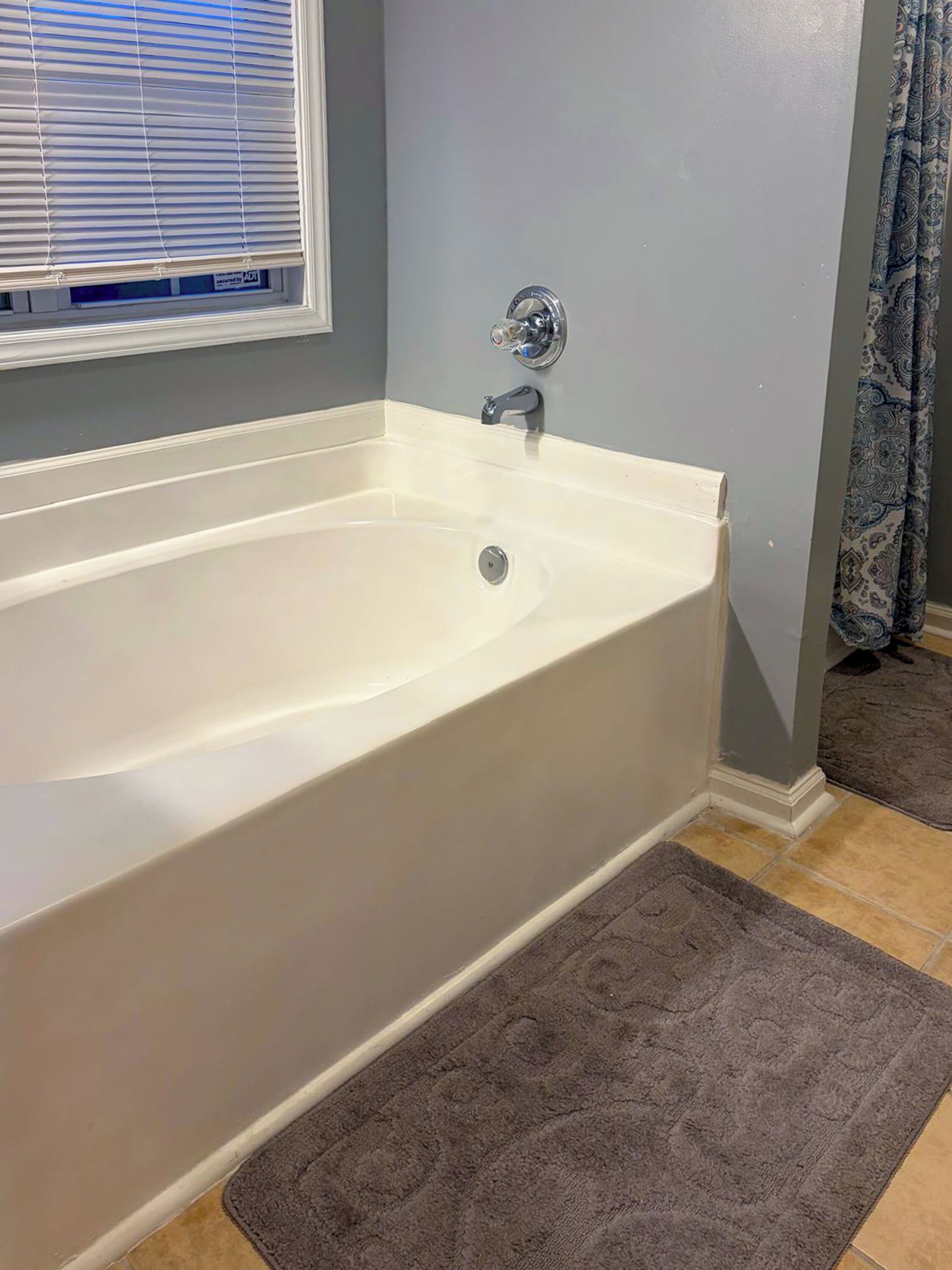A bathroom with a white bathtub, a window with blinds, and a patterned curtain.
