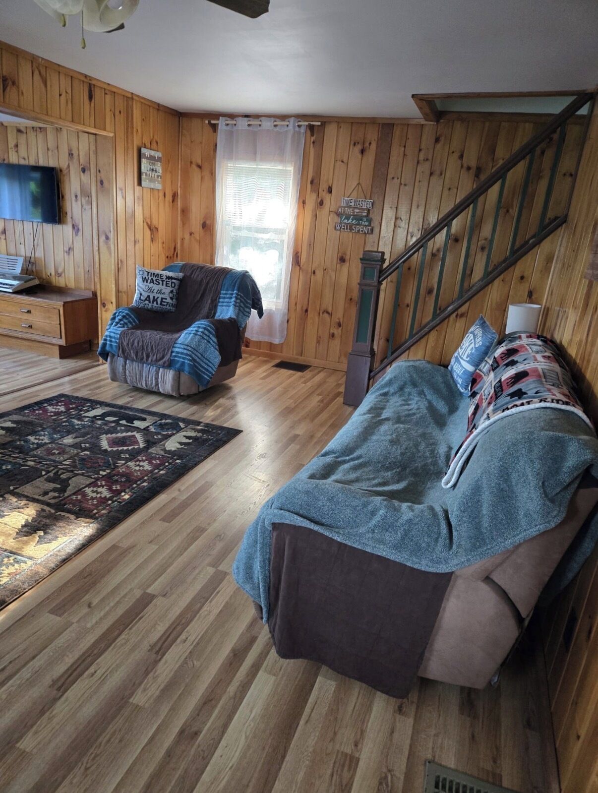 A room with wooden walls, a bed with a blanket, a chair with a cushion, a small table, and a television.