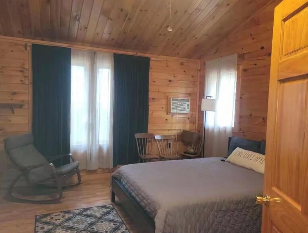 Quarto com painéis de madeira, uma cama, duas cadeiras, uma mesinha e um abajur.