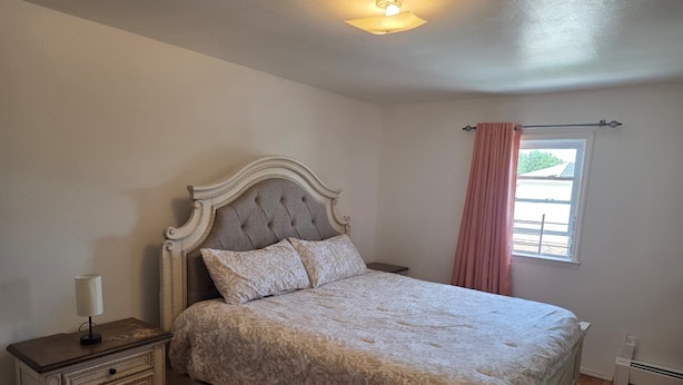Quarto com cama, mesa de cabeceira, luminária, janela com cortinas e luz no teto.