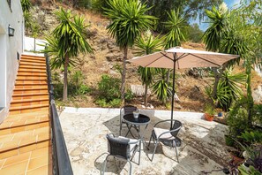 Eine Terrasse mit Gartenmöbeln, einem Tisch und Stühlen, umgeben von Pflanzen und Blick auf eine Hügellandschaft.