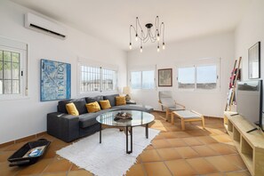 Un salón amplio con un sofá seccional gris, una mesa de centro de vidrio y una silla de madera. Amplias ventanas ofrecen luz natural y vistas al exterior.
