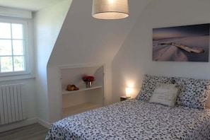 A bedroom with a bed, a shelf with flowers, a painting, and a window.