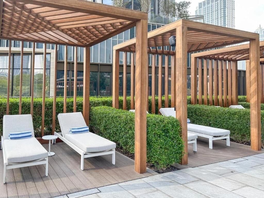 A rooftop lounge area with wooden pergolas, white lounge chairs, and a neatly trimmed hedge.