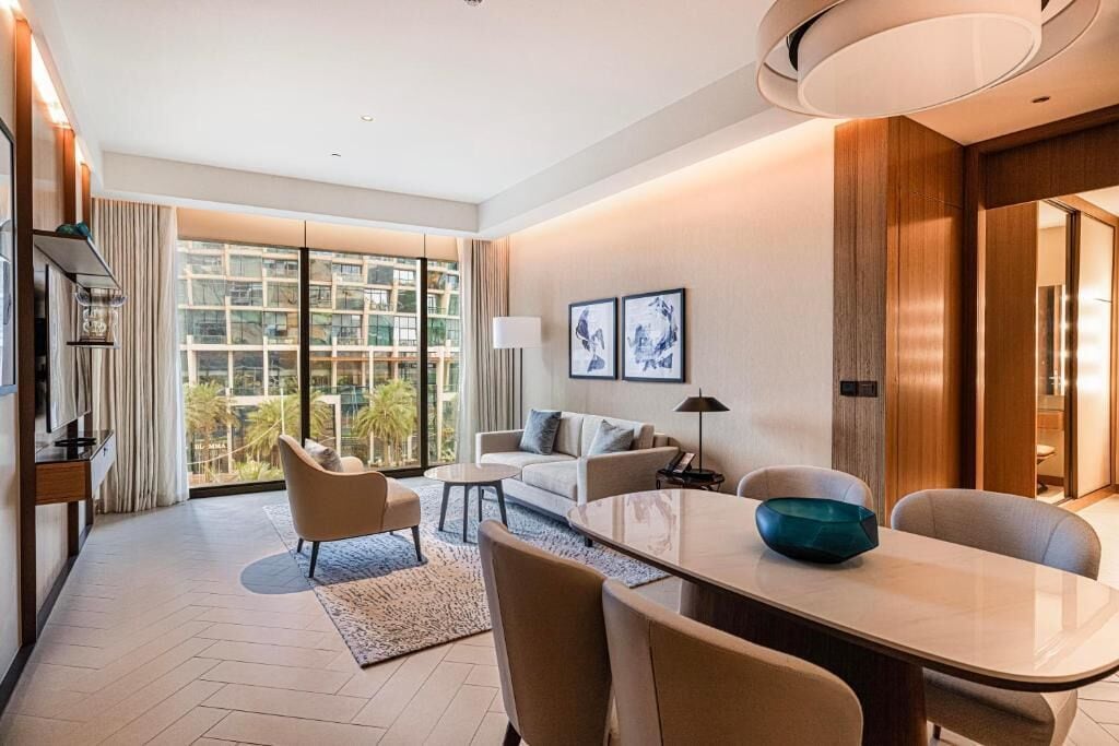 A modern hotel room with a large window, a sofa, a dining table, and a lamp.