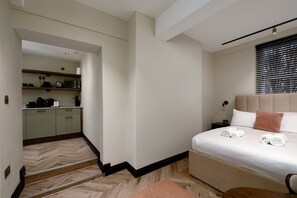 A hotel room with a bed, a nightstand, a window with blinds, and a view of the kitchen area. - GuestReady - Stylish studio in Euston (London)