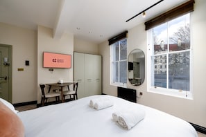 Ein Hotelzimmer mit Bett, Schreibtisch, Sessel, Fernseher und einem Fenster mit Blick auf Gebäude.